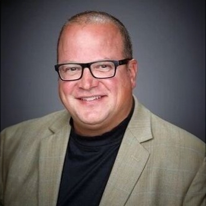 A man wearing a light-colored blazer and glasses, smiling at the camera.