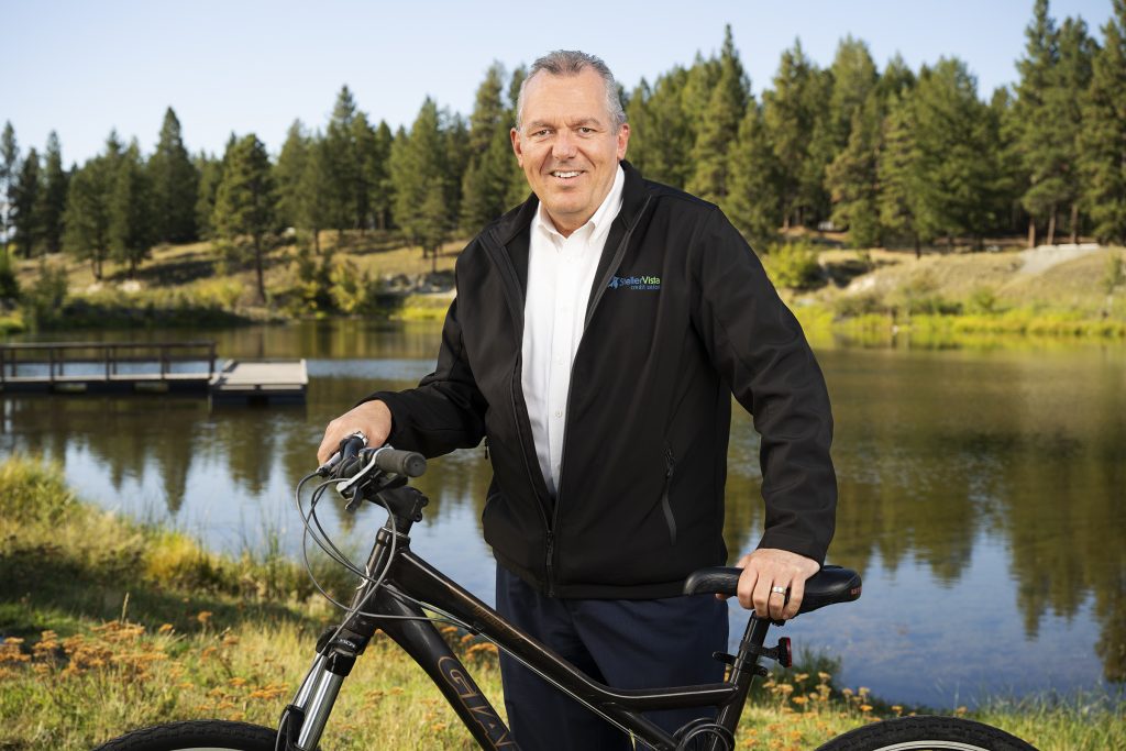 Man in a black jacket standing next to a bicycle by a lake surrounded by trees.
