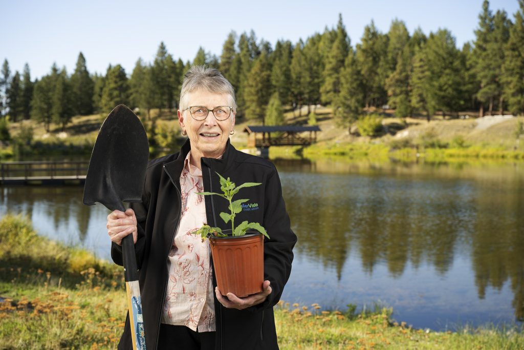 Woman with glasses holding a potted plant and a shovel near a lake surrounded by trees.