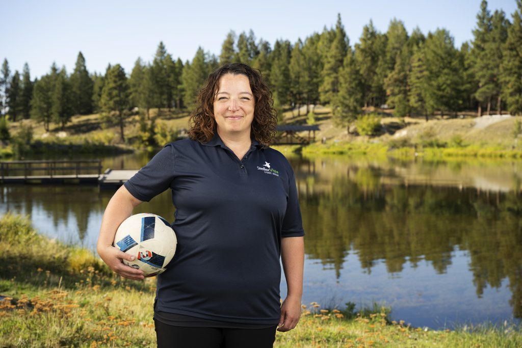 Woman in a navy polo shirt holding a soccer ball while standing by a lake surrounded by trees.