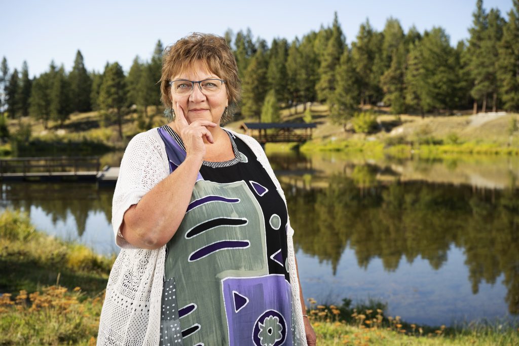Woman with glasses wearing a patterned dress standing by a lake surrounded by trees.