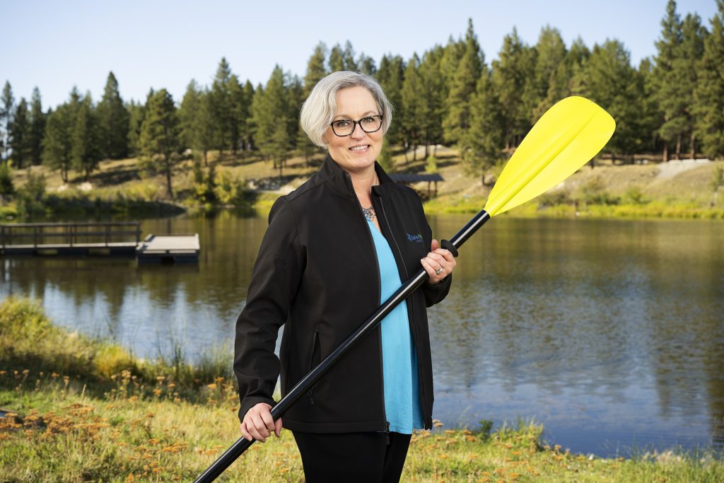 Woman with short gray hair holding a yellow kayak paddle near a lake surrounded by trees.