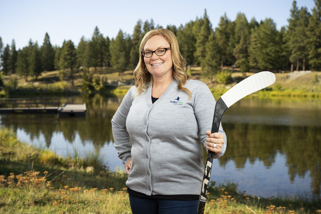 Woman with glasses holding a hockey stick while standing by a lake surrounded by trees.