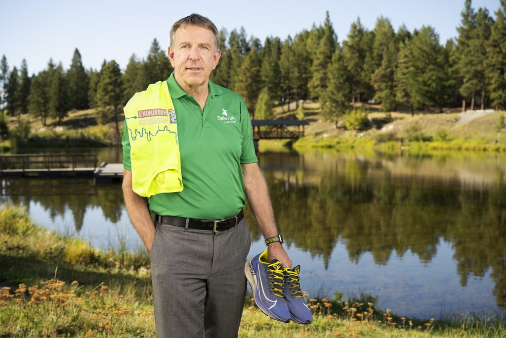 Man in a green shirt holding running shoes while standing by a lake with trees in the background.